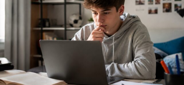 Teen studying at a desk with a laptop, open book, and notebook, illustrating the use of AI tools like ChatGPT for summer reading and school assignments.