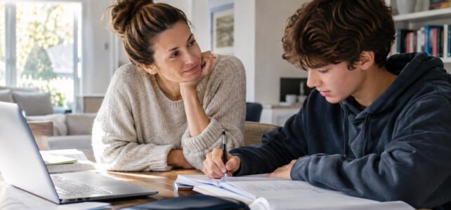 Mother sitting beside her teenage son as he works on homework at a kitchen table with books and a laptop.